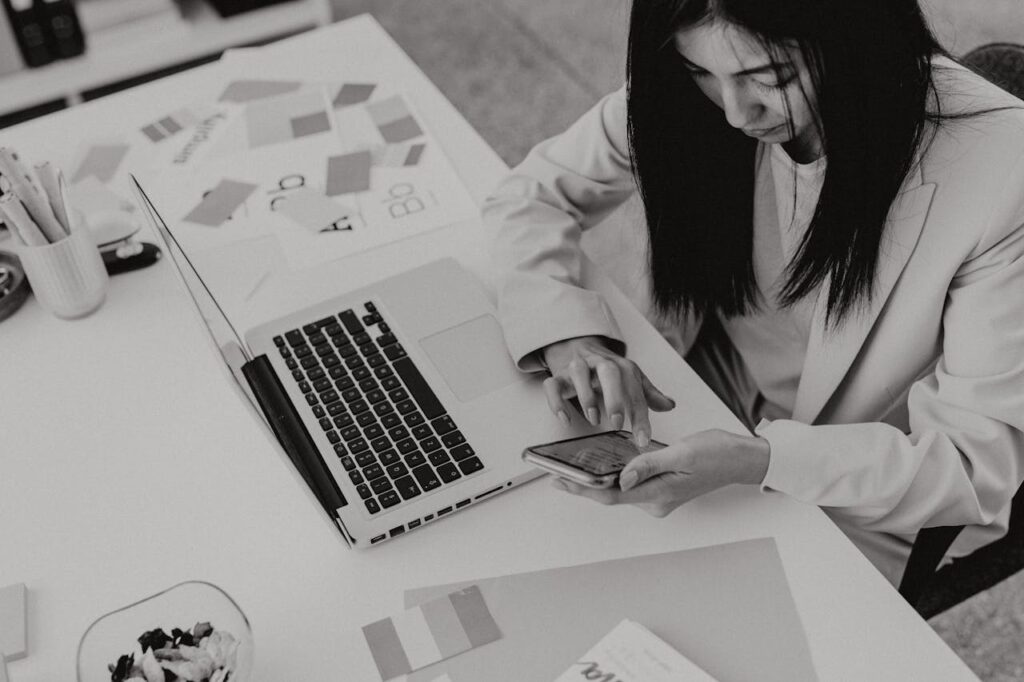 Asian businesswoman in office, using smartphone while working at laptop in black and white setting.